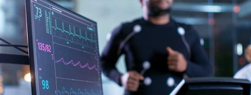 Close-up Shot of a Monitor With EKG Data. Male Athlete Runs on a Treadmill with Electrodes Attached to His Body while Sport Scientist Holds Tablet and Supervises EKG Status in the Background. - Colorado Center of Orthopaedic Excellence sports performance